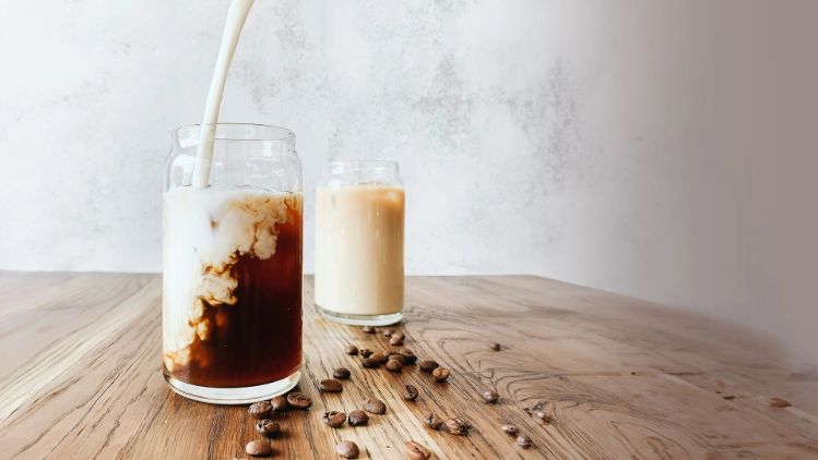 Vaso de café frío sobre una mesa de madera mientras se le vierte leche, creando remolinos claros en el café oscuro. Al fondo, otro vaso con café con leche y granos de café dispersos sobre la superficie.